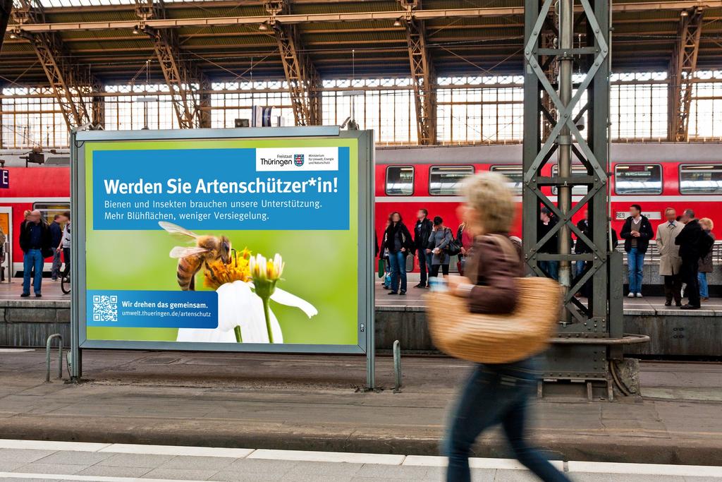 Plakat auf einem Bahnhof mit vielen Menschen, Aufschrift neben Biene auf einer Blüte: Werden Sie Artenschützer