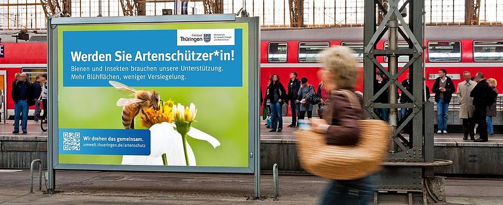 Werden Sie Artenschützer! Plakat auf einem Bahnhof mit vielen Menschen, Aufschrift neben Biene auf einer Blüte: Werden Sie Artenschützer
